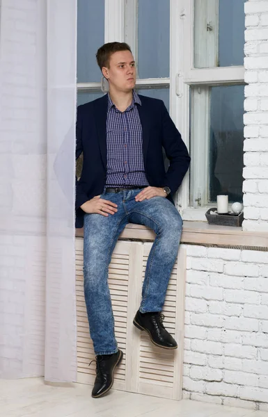 Jeune homme avec une expression arrogante. Assis près de la fenêtre sur le rebord de la fenêtre . — Photo