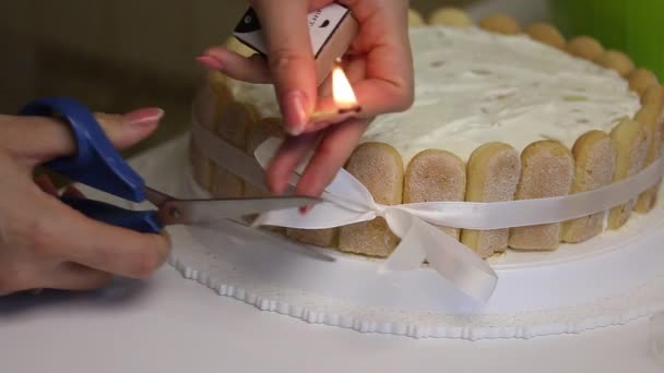 Op tafel ligt een taart van Savoiardi koekjes, gebonden met een lint met strik. Een vrouw smelt de uiteinden van een lint met lucifers. — Stockvideo