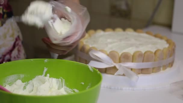 Sul tavolo c'è una torta di biscotti Savoiardi, legati con un nastro con un fiocco. Donna mette la crema in una borsa da pasticceria . — Video Stock