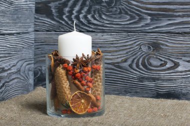 Cam bir kapta büyük bir mum. Koniler, dağ külleri, anason yıldızları, tarçın çubukları dekorasyon için içine dökülüyor. Fırçalanmış tahtaların arkasında duruyor..