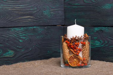 Cam bir kapta büyük bir mum. Koniler, dağ külleri, anason yıldızları, tarçın çubukları dekorasyon için içine dökülüyor. Fırçalanmış tahtaların arkasında duruyor..