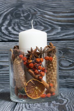 Cam bir kapta büyük bir mum. Koniler, dağ külleri, anason yıldızları, tarçın çubukları dekorasyon için içine dökülüyor. Siyah beyaz boyanmış tahtalarda duruyor..
