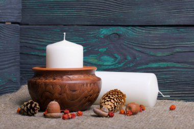 Kil bir kapta büyük bir mum. Meşe palamutları, Rowan çilekleri yakınlarda ve bir mum uzanıyor. Fırçalanmış tahtaların arka planına karşı.