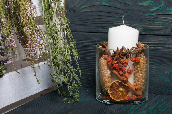 A large candle in a glass vessel. Cones, mountain ash, anise stars, cinnamon sticks are poured into it for decoration. Bunches of fragrant herbs hang nearby. It stands on painted boards painted in bla