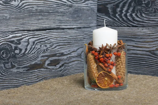 A large candle in a glass vessel. Cones, mountain ash, anise stars, cinnamon sticks are poured into it for decoration. It stands on the background of brushed boards.