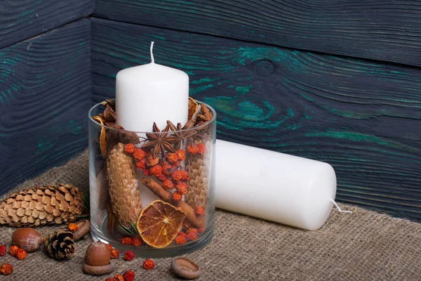 A large candle in a glass vessel. Cones, mountain ash, anise stars, cinnamon sticks are poured into it for decoration. Acorns, rowan berries are scattered nearby and a candle lies. Against the backgro