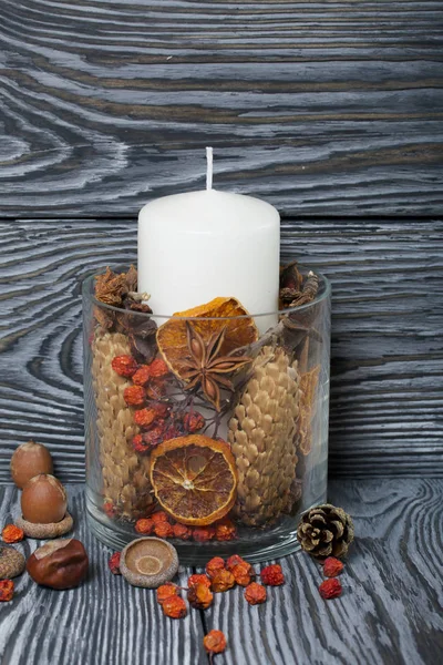 Una grande candela in un vaso di vetro. Coni, cenere di montagna, stelle di anice, bastoncini di cannella sono versati in esso per la decorazione. Le ghiande e le ceneri di montagna sono sparse nelle vicinanze. Cavalletti su tavole dipinte dipinte in — Foto Stock