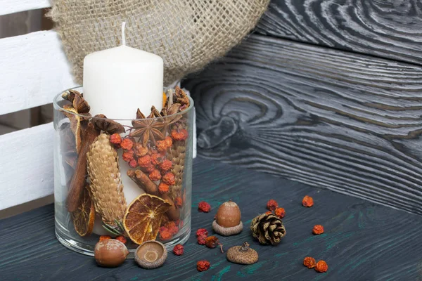 A large candle in a glass vessel. Cones, mountain ash, anise stars, cinnamon sticks are poured into it for decoration. Stands on brushed. — Stock Photo, Image