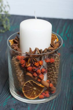 Cam bir kapta büyük bir mum. Koniler, dağ külleri, anason yıldızları, tarçın çubukları dekorasyon için içine dökülüyor. Siyah ve yeşile boyanmış tahtaların üzerinde duruyor..
