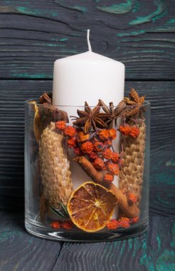 Cam bir kapta büyük bir mum. Koniler, dağ külleri, anason yıldızları, tarçın çubukları dekorasyon için içine dökülüyor. Siyah ve yeşile boyanmış tahtaların üzerinde duruyor..