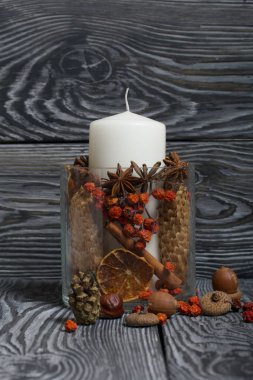 Cam bir kapta büyük bir mum. Koniler, dağ külleri, anason yıldızları, tarçın çubukları dekorasyon için içine dökülüyor. Meşe palamudu ve dağ külleri etrafa saçılmış. Boyalı tahtaların üzerinde durur