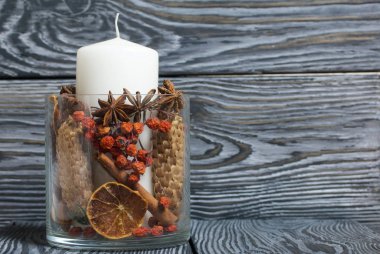 Cam bir kapta büyük bir mum. Koniler, dağ külleri, anason yıldızları, tarçın çubukları dekorasyon için içine dökülüyor. Siyah beyaz boyanmış tahtalarda duruyor..