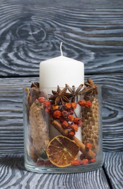 Cam bir kapta büyük bir mum. Koniler, dağ külleri, anason yıldızları, tarçın çubukları dekorasyon için içine dökülüyor. Siyah beyaz boyanmış tahtalarda duruyor..