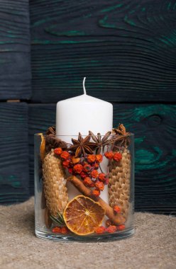 Cam bir kapta büyük bir mum. Koniler, dağ külleri, anason yıldızları, tarçın çubukları dekorasyon için içine dökülüyor. Fırçalanmış tahtaların arkasında duruyor..
