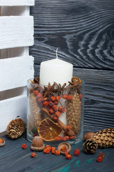 A large candle in a glass vessel. Cones, mountain ash, anise stars, cinnamon sticks are poured into it for decoration. Acorns and mountain ash are scattered nearby. Stands on painted boards painted in