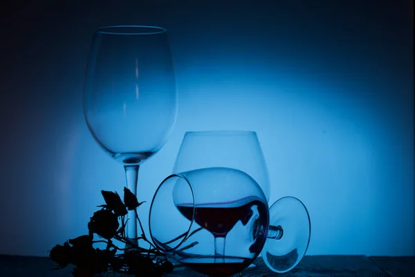 Glasses with red dry wine. One of them lies on its side with the remains of wine. Dried rose flowers. Stand on wooden boards. Shot in backlight. — Stock Photo, Image