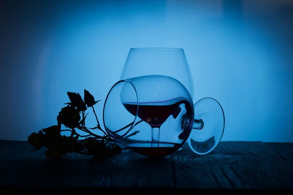 Okulary z czerwonym suchym winem. Jeden z nich leży po swojej stronie z pozostałościami wina. Suszone kwiaty róż. Stań na drewnianych deskach. Strzał w podświetlenie. — Zdjęcie stockowe