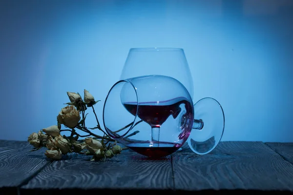 Sklenice s červeným suchým vínem. Jeden z nich leží na boku s pozůstatky vína. Sušené květy růží. Postavte se na dřevěné desky. Výstřel v podsvícení. — Stock fotografie