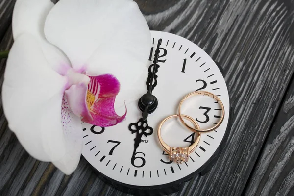 Весільні кільця та квітка орхідеї знаходяться на годиннику. На матованих соснових дошках пофарбованих у чорно-білий колір . — стокове фото