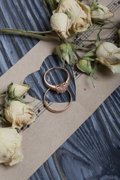 Los anillos de boda se encuentran sobre el papel con el corazón cortado. Rosas secas cercanas. Sobre tablas de pino cepillado pintadas en blanco y negro . —  Fotos de Stock