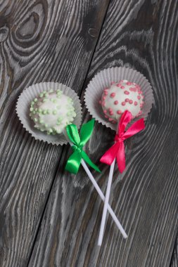 Cake pops in white glazed chocolate with green and pink sprinkles. A green and pink bow is tied on sticks. Against the background of brushed pine boards painted in black and white.