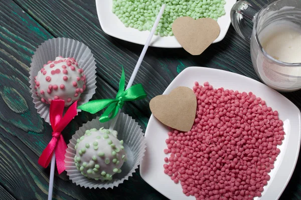 Cake pops in white glazed chocolate with green and pink sprinkles. A green and pink bow is tied on sticks. Near topping in plates and hearts of paper. Against the background of brushed pine boards pai