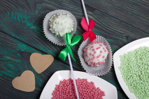 Cake pops in white glazed chocolate with green and pink sprinkles. A green and pink bow is tied on sticks. Near topping in plates and hearts of paper. Against the background of brushed pine boards pai