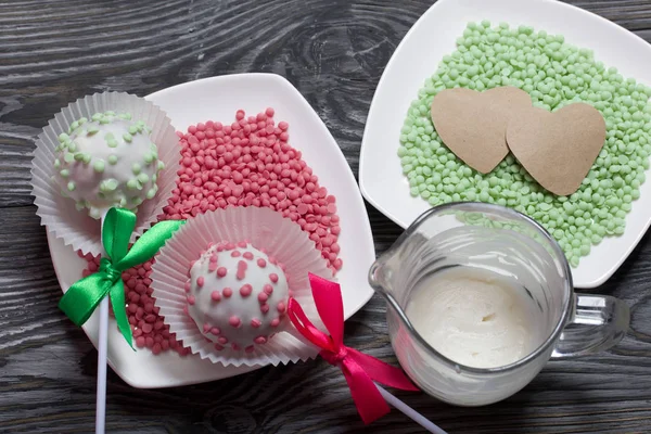 Cake pops in white glazed chocolate with green and pink sprinkles. A green and pink bow is tied on sticks. Nearby melted chocolate and topping in plates. Against the background of brushed pine boards