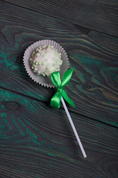Cake pops in white glazed chocolate with green and pink sprinkles. A green and pink bow is tied on sticks. Against the background of brushed pine boards painted in black and white.