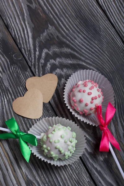 Cake pops in white glazed chocolate with green and pink sprinkles. A green and pink bow is tied on sticks. Nearby hearts cut out of paper. Against the background of brushed pine boards painted in blac