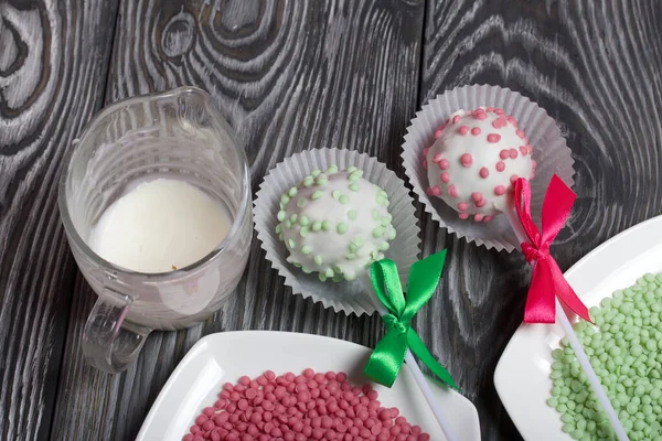 Cake pops in white glazed chocolate with green and pink sprinkles. A green and pink bow is tied on sticks. Nearby melted chocolate and topping in plates. Against the background of brushed pine boards — Stock Photo, Image