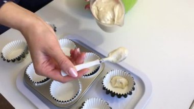 A woman puts dough in cupcake baking dishes. Nearby are empty forms for filling.