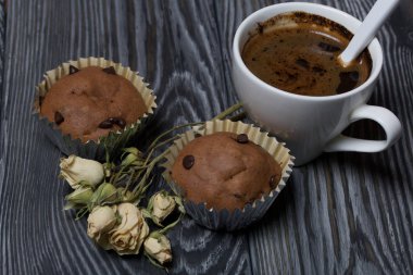 Beyaz bardakta kahve. Yakınlarda çörekler ve kurutulmuş bej rengi bir gül var. Çam ağacından yapılmış tahtalarda.