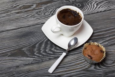 Beyaz fincanda kahve bir fincan tabağında duruyor. Yakınlarda bir kek ve çay tabağında bir kaşık var. Çam ağacından yapılmış tahtalarda.