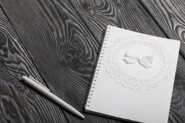 Not defteri beyazı ve spiral. Kapağı kağıt dantel ve kurdeleyle süslenmiş. Yakınlarda beyaz bir kalem var. Çam ağacından yapılmış tahtalarda.