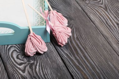 Çubukta kalp şeklinde narin pembe şekerlemeler. Bir tanesi kurdeleyle fiyonk bağlanmış. Turkuaz renkli bir tepside yatıyor. Taranmış çam ağaçlarının arka planında siyaha boyanmış.