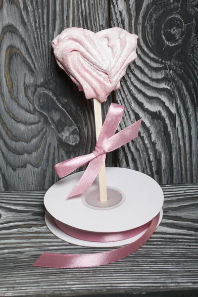 Jemné růžové marshmallows ve tvaru srdce na klacku. S mašlí přivázanou k luku. Poblíž cívky s copem. Leží na kartáčovaných borovicových prknech malovaných v černé a bílé. — Stock fotografie