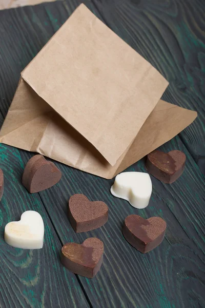 An open white paper bag on brushed pine boards. Chocolate hearts are scattered from it. From white and black chocolate.