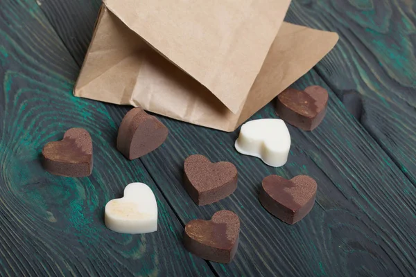 An open white paper bag on brushed pine boards. Chocolate hearts are scattered from it. From white and black chocolate.
