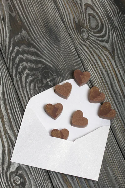 An open white paper envelope on brushed pine boards. Chocolate hearts are scattered from it. — Stock Photo, Image