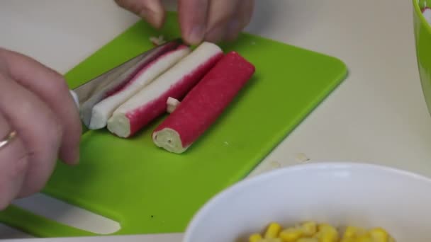 Un hombre hace una ensalada. Rebanadas de palitos de cangrejo en una tabla de cortar y se transfiere a un recipiente. En primer plano hay un contenedor de maíz. Primer plano de disparo — Vídeo de stock