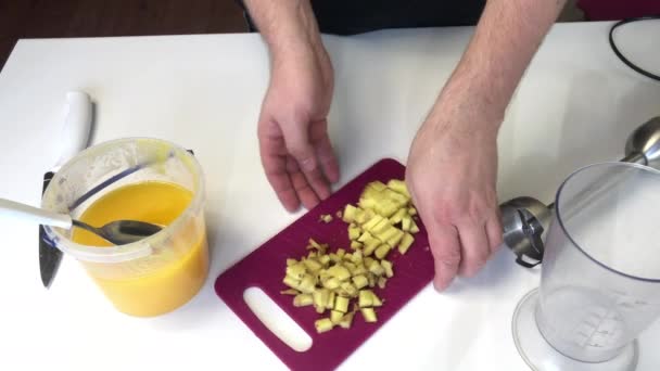 Un homme prend une planche à découper avec du gingembre haché et la verse dans un bol de mélangeur. A proximité se trouve du miel dans un récipient et les éléments du mélangeur sont visibles. Pour cuire le gingembre au miel . — Video