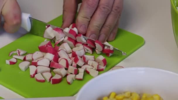 Un hombre hace una ensalada. Rebanadas de palitos de cangrejo en una tabla de cortar y se transfiere a un recipiente. En primer plano hay un contenedor de maíz. Primer plano de disparo . — Vídeos de Stock