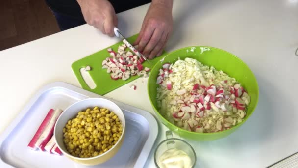 A man makes a salad. Slices crab sticks on a cutting board and adds to a container with chopped Beijing cabbage. In other containers, corn and mayonnaise — Stock Video