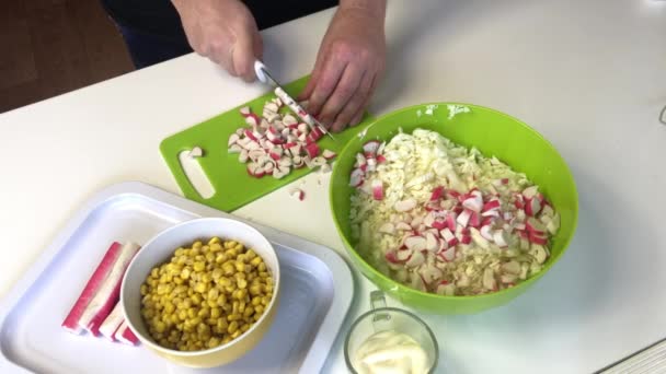 En man gör en sallad. Skivor krabba pinnar på en skärbräda och lägger till en behållare med hackad Peking kål. I andra behållare: majs och majonnäs — Stockvideo