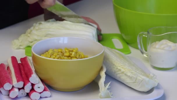 Um homem faz uma salada de couve de Pequim. Repolho de partes com uma faca em uma tábua de redução. Ao lado da mesa estão outros ingredientes. Milho, paus de caranguejo e maionese. Imagem de close-up — Vídeo de Stock