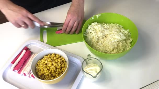 Ein Mann macht einen Salat. Krabbenstäbchen auf einem Schneidebrett schneiden. Neben dem Container liegt geschredderter Chinakohl. Mais und Mayonnaise in Containern. — Stockvideo