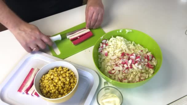 Ein Mann macht einen Salat. Krabbenstäbchen auf einem Schneidebrett schneiden. Neben dem Container liegt geschredderter Chinakohl. Mais und Mayonnaise in Containern — Stockvideo