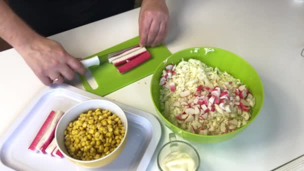 Un hombre hace una ensalada. Rebanadas de palitos de cangrejo en una tabla de cortar. Junto al contenedor se desmenuza la col de Beijing. Maíz y mayonesa están en recipientes — Vídeos de Stock