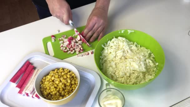 Seorang pria membuat salad. Mengiris batang kepiting di papan potong dan menambahkan ke wadah dengan potongan kubis Beijing. Dalam wadah lain, jagung dan mayones . — Stok Video